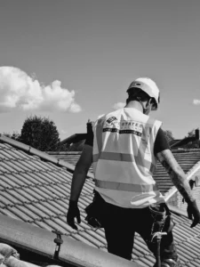 Black and white image of a professional roofer wearing an Estates Roofing high-visibility vest and safety gear works on a tiled roof under a bright sky, representing expert roofing services